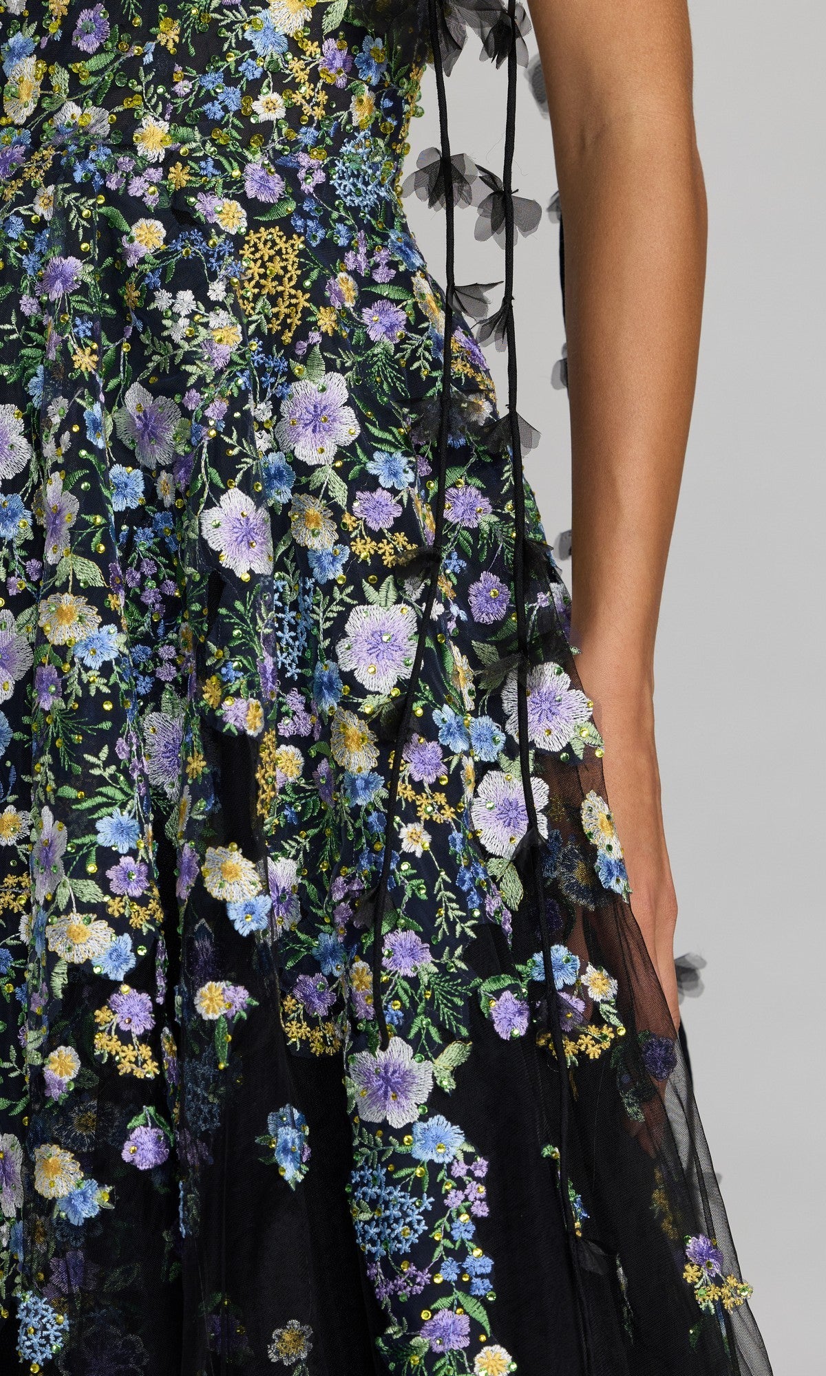 Close-up of a person wearing the Midi Party Dress: Mac Duggal 21356—a black tulle midi dress featuring blue, purple, and yellow floral embroidery in an elegant A-line silhouette—with their right arm resting at their side.